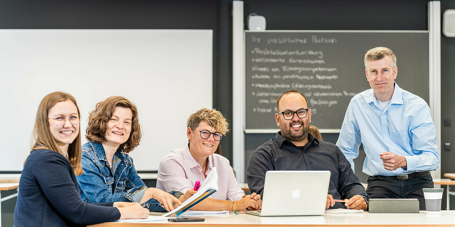 Studierende und ihr Dozent arbeiten mit ihren Lernmaterialien an einem Tisch.