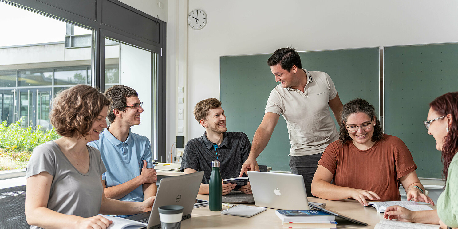 Studierende arbeiten in einer Lerngruppe mit Laptops und Unterlagen an einem großen Tisch.