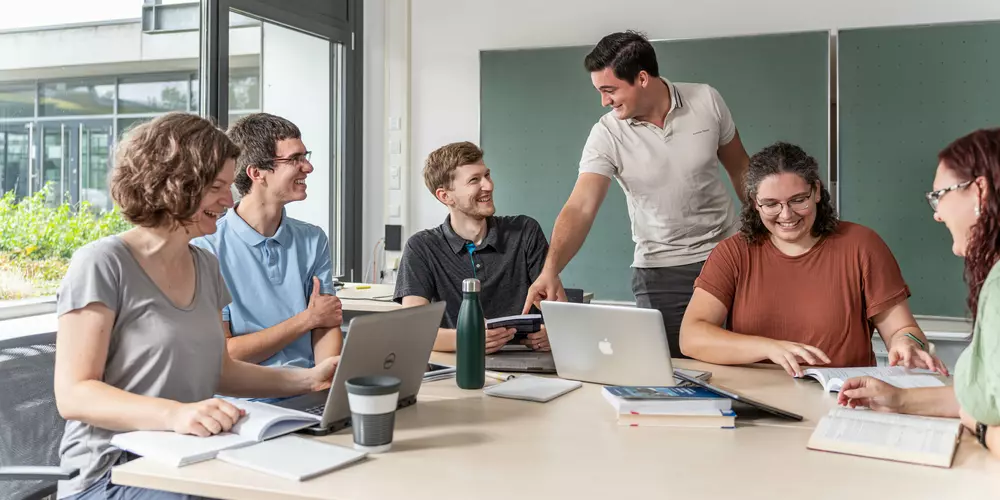 Studierende arbeiten in einer Lerngruppe mit Laptops und Unterlagen an einem großen Tisch.