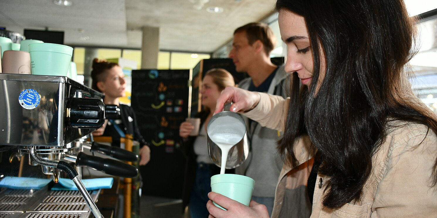 Eine Frau gießt Milch in einen Kaffeebecher. Im Hintergrund unterhalten sich Menschen.