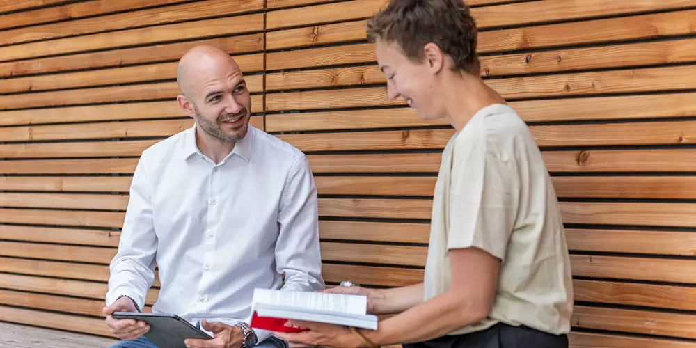 Zwei Studierende sitzen mit Büchern auf einer Bank in einer Holznische.