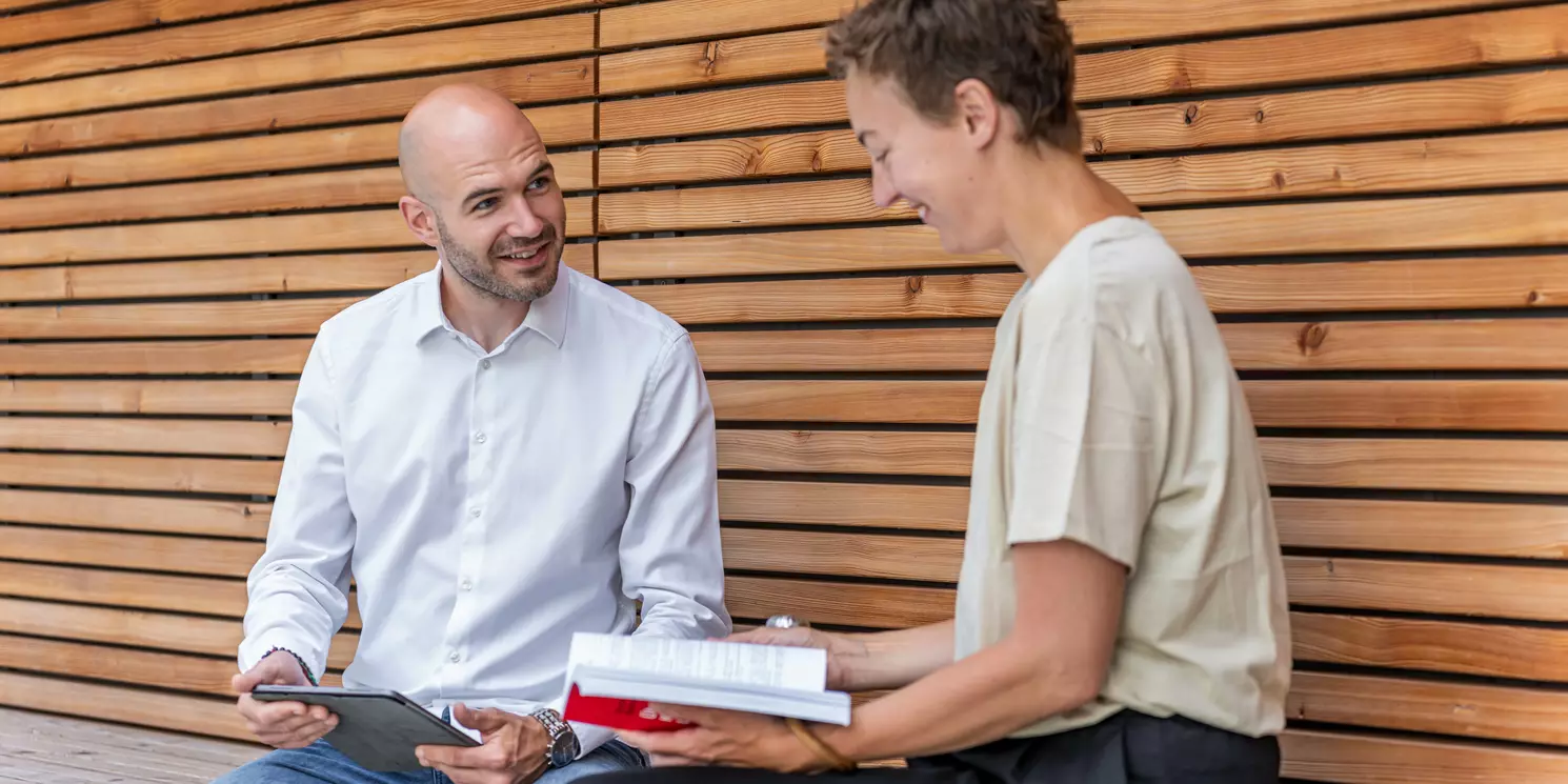 Zwei Studierende sitzen mit Büchern auf einer Bank in einer Holznische.