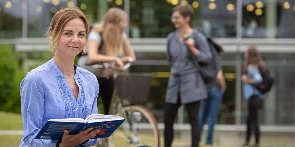 Studentin sitzt mit einem Buch vor der Bibliothek. Im Hintergrund unterhalten sich Studierende.